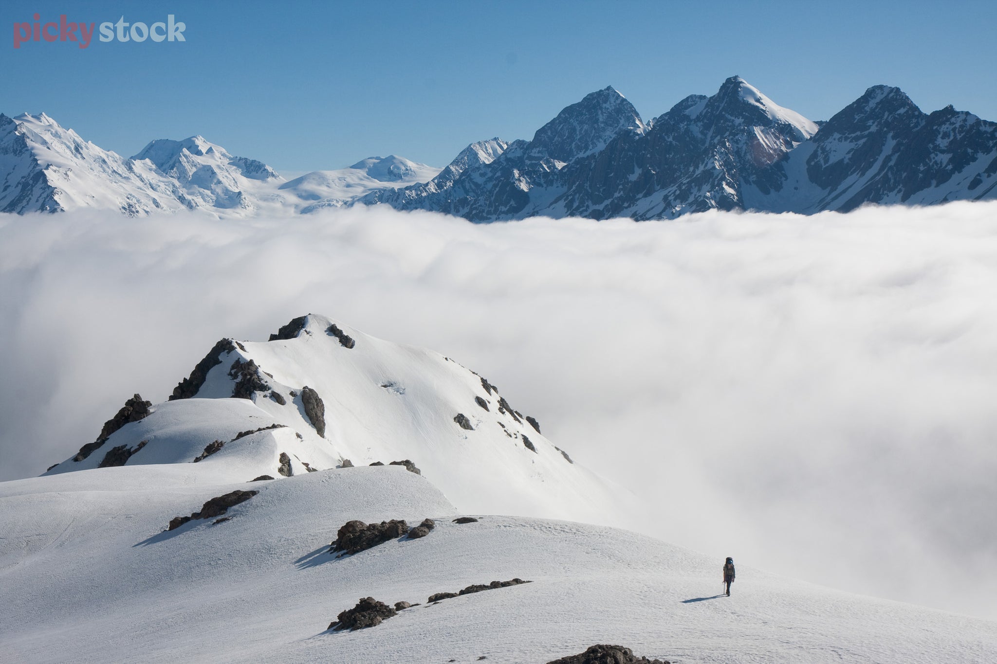 Lone tramper sits dwarfed by a snowy mountain as they walk the ridgeline above cloud level. Mountain peaks just pop above the clouds in the far distance. 