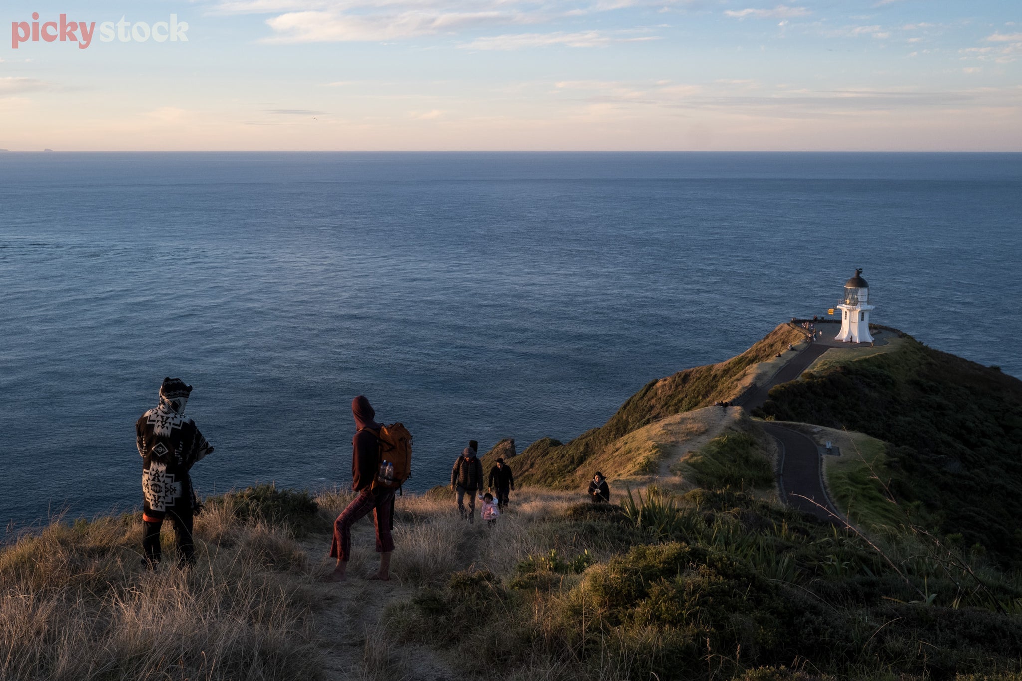 Line of people on walking track up the hill from Cape Reinga Lighthouse, looking back down towards the white lighthouse with sea in the background. 
