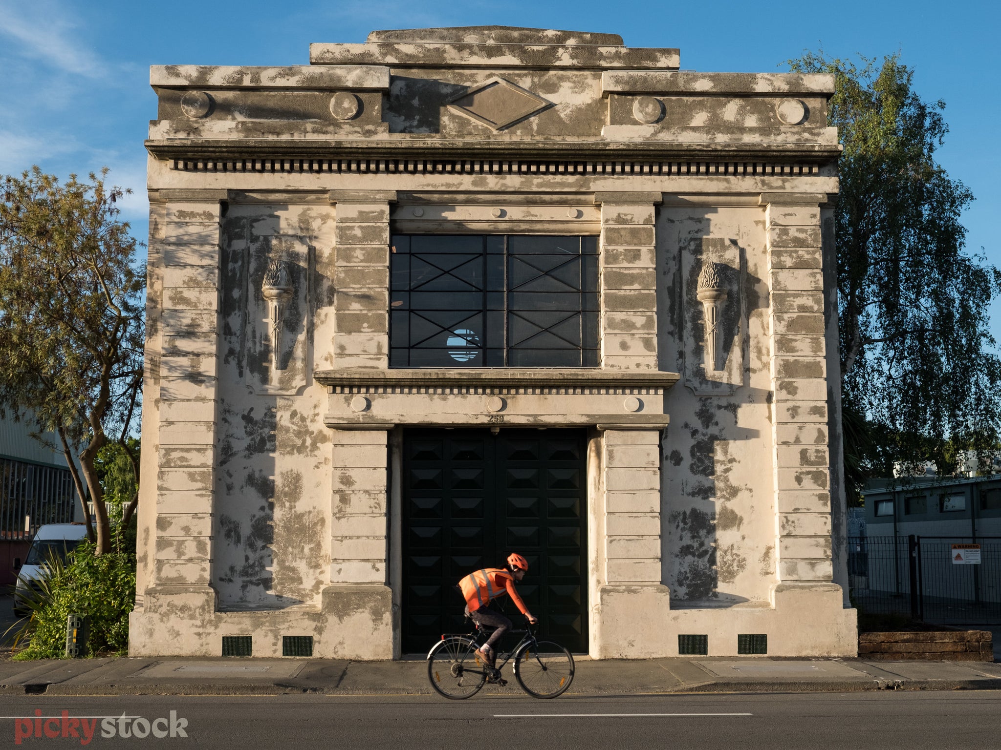 Cyclist with hi-vis vest, fluro orange helmet and socks rides past old building on a blue day. 