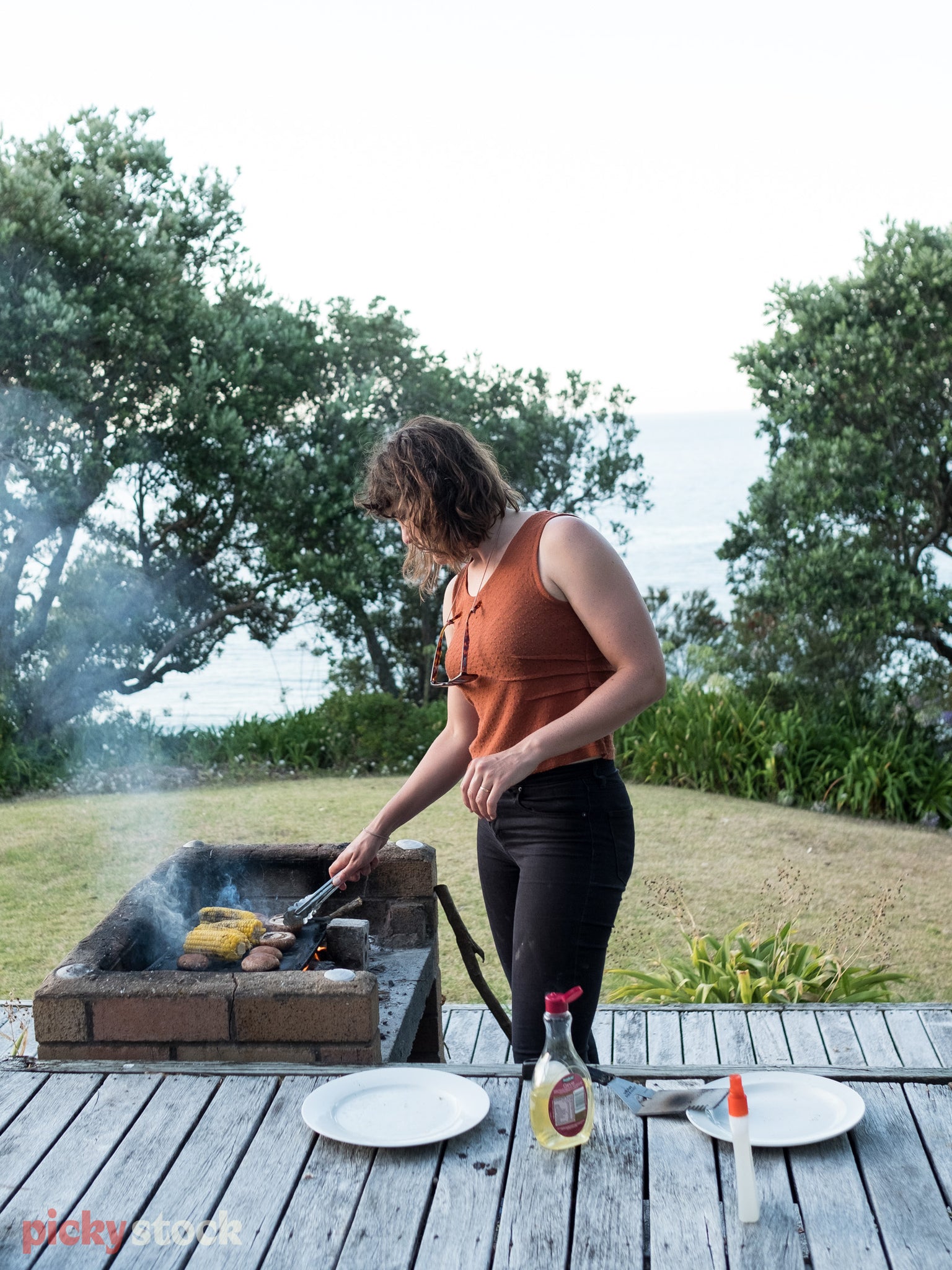 Red head lady in early twenties cooks on old charcoal BBQ, getting it heated and ready for cooking. She wears a burnt umbre coloured singlet and her hair is semi-wet. 