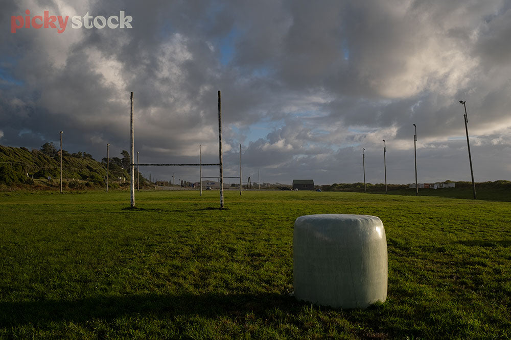 Single hay bale sitting on large rugby sports field. Goal posts in the background. Low cloud, dark sky, early evening. 