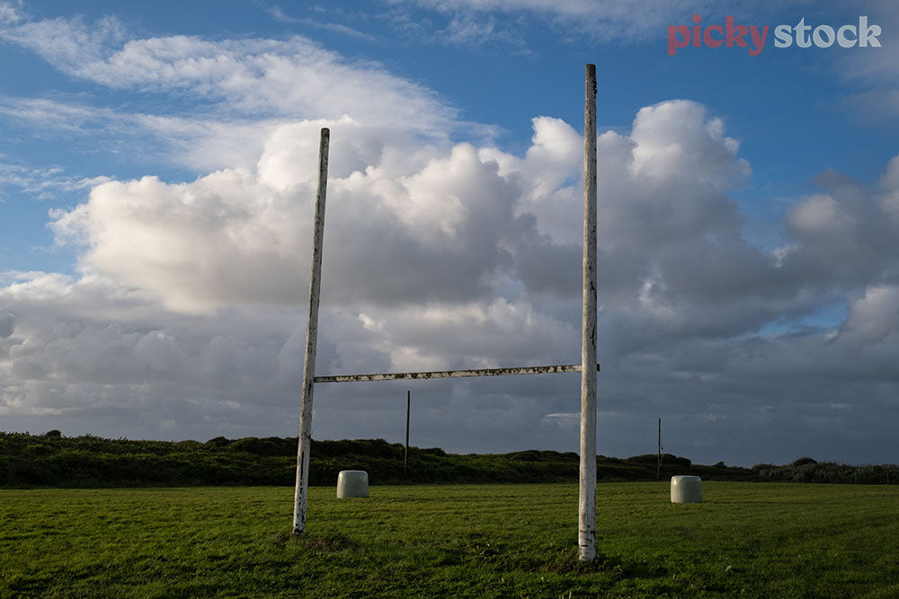 Rugby posts on a rural green grasses field. Poles are worn and exposed. Hay bales in the background marking either side of the post. Sky is blue with thick cloud. 