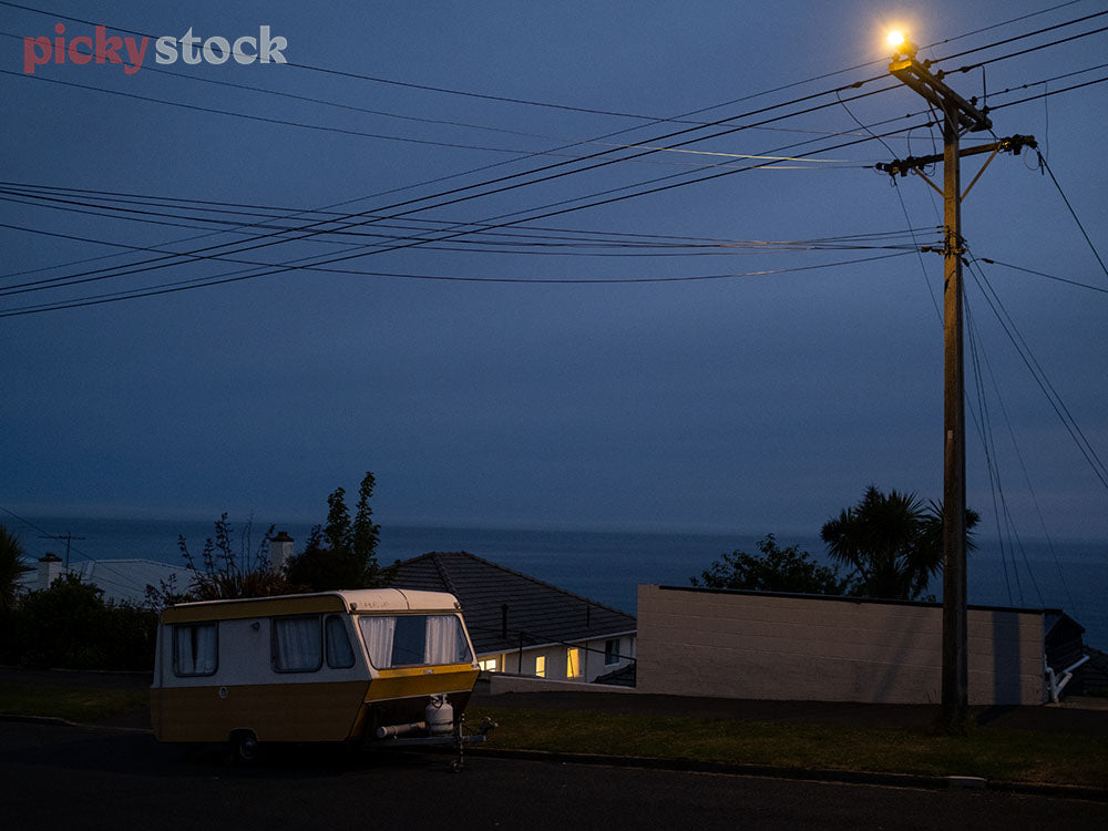 Evening dark blue sky. Power lines with large pole to the right of frame, turned on. Soft yellow light. Caravan yellow on side of the road under the light. Houses in the background with ocean view. 