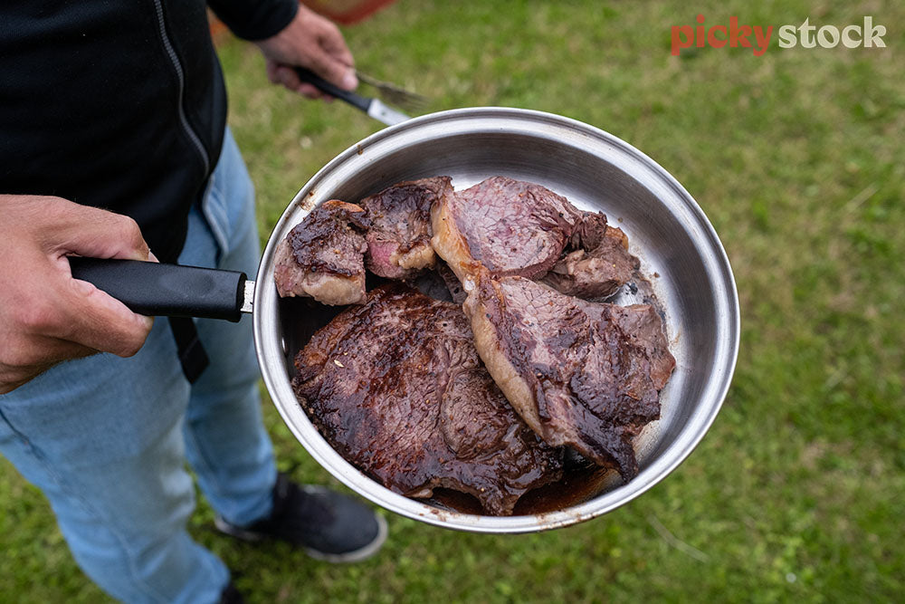Man in black top and blue jeans holding a fry pan with a pile of cooked steaks. Holding a knife in the other hand