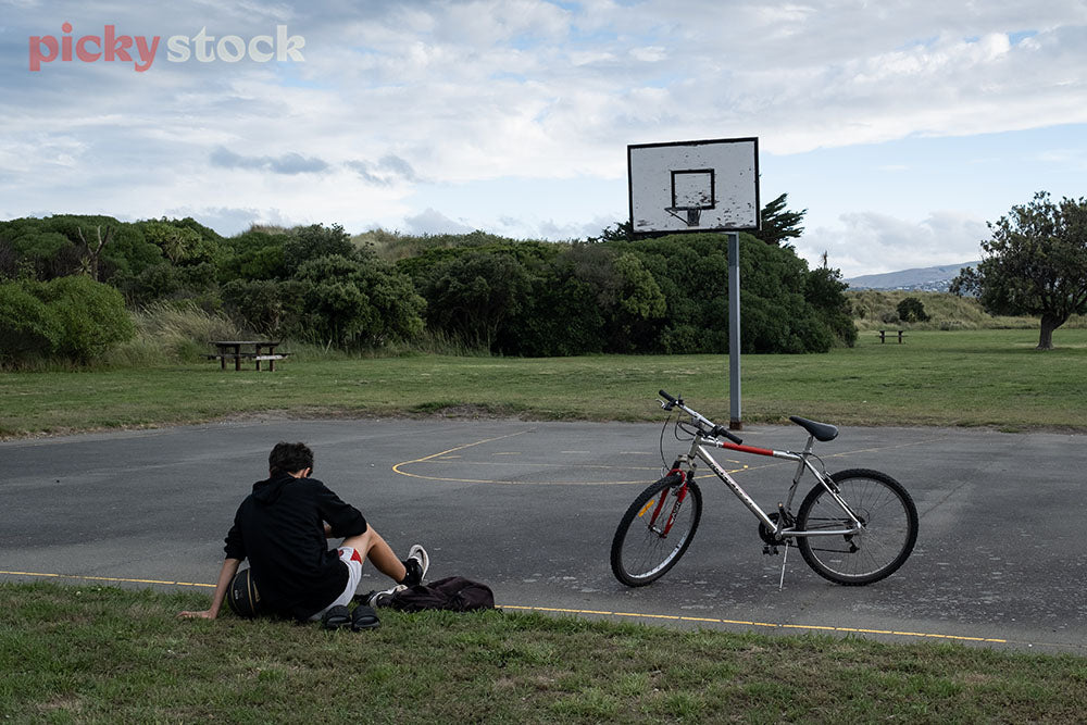 Young kid sitting on grass on basketball court boundary. Push bike standing parked on concrete court. Green park and field surrounding him.