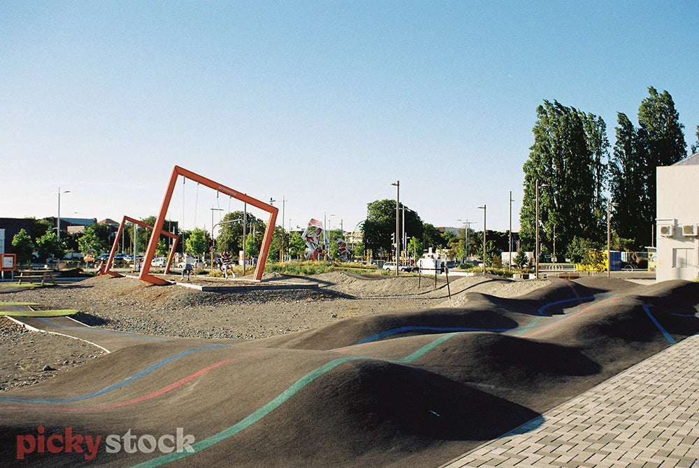 A pump track for bikes and skateboards in Central Christchurch. In the background are large orange square frames and two people swinging on swings underneath them. Behind that are trees and street lamps.