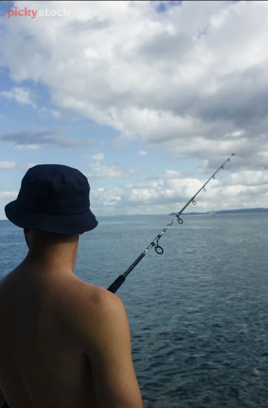 Looking out over the shoulder of a bucket-hat-wearing man fishing. His shirtless body takes up the left hand side of frame, while his fishing rod extends out to the right. The sea meets the horizon halfway up the image. 