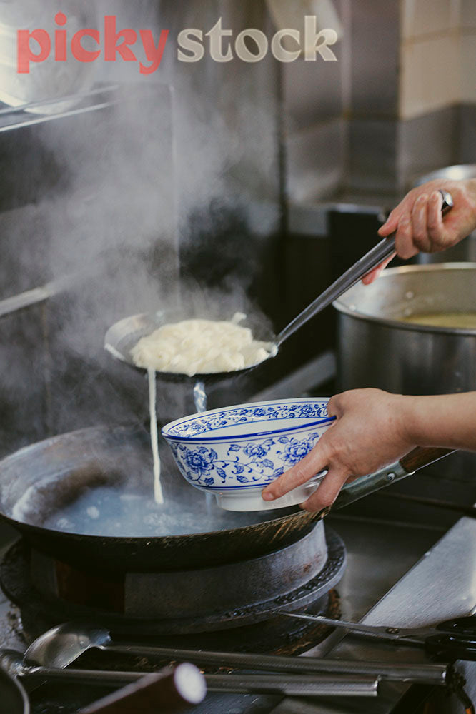 Chef ladles noodes into big bowl over steaming wok. 