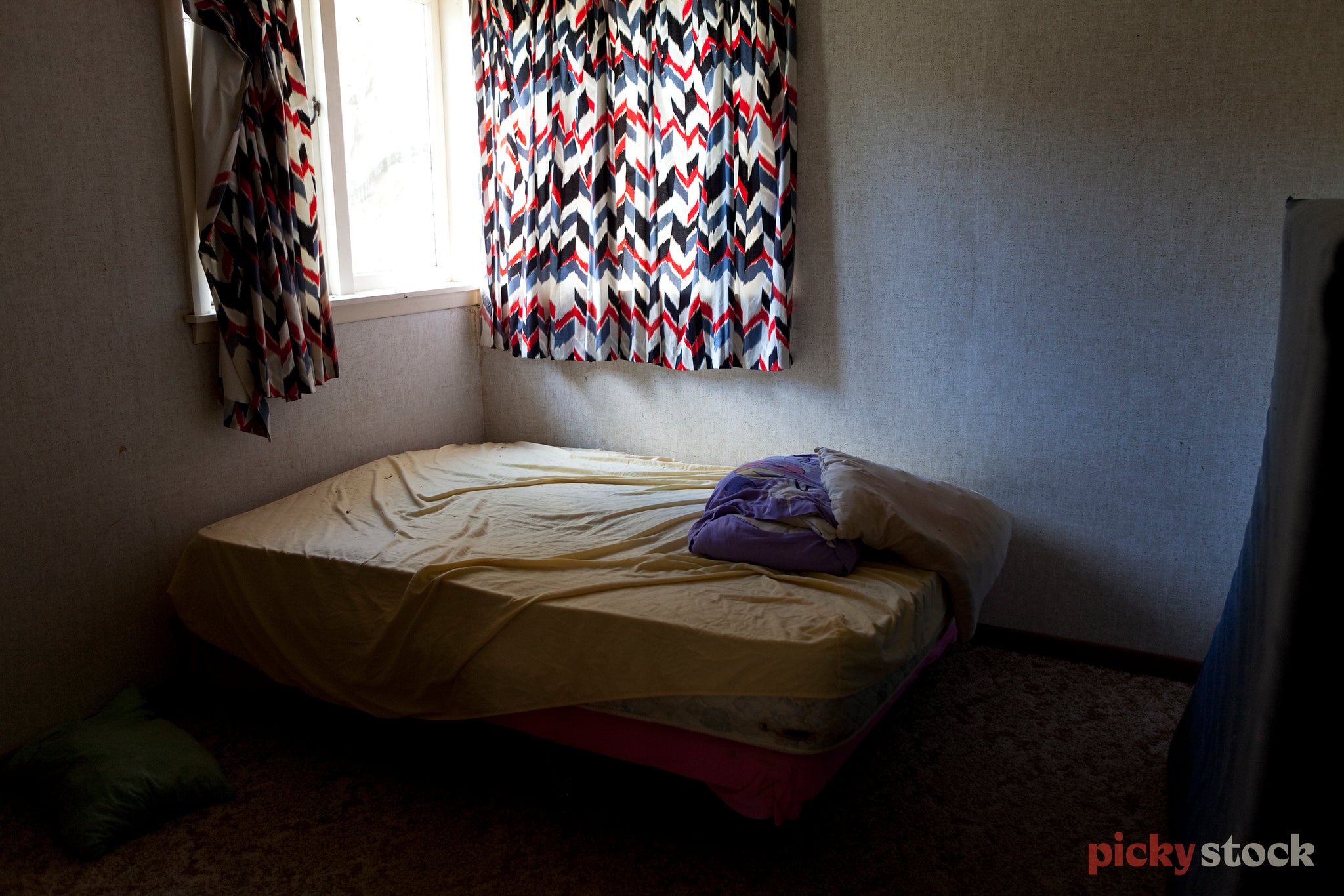 A empty unmade bed with old sheets and an old duvet sit in a silent room.The half open, patterned curtains bring much needed light into the room. 