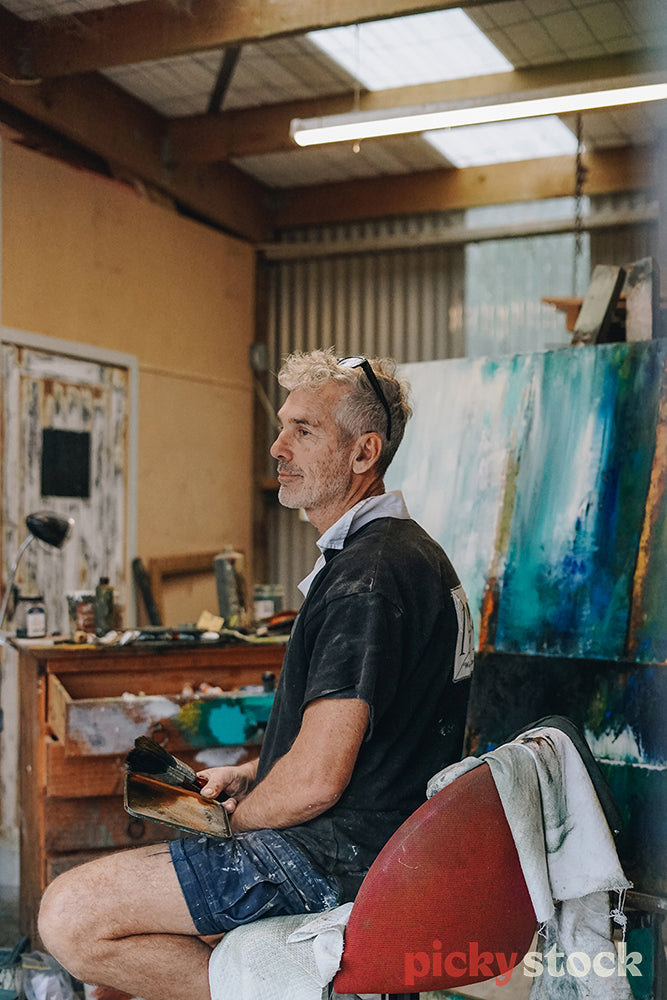 Painter in old rugby jersey sits in his studio, gazing off into distance. 