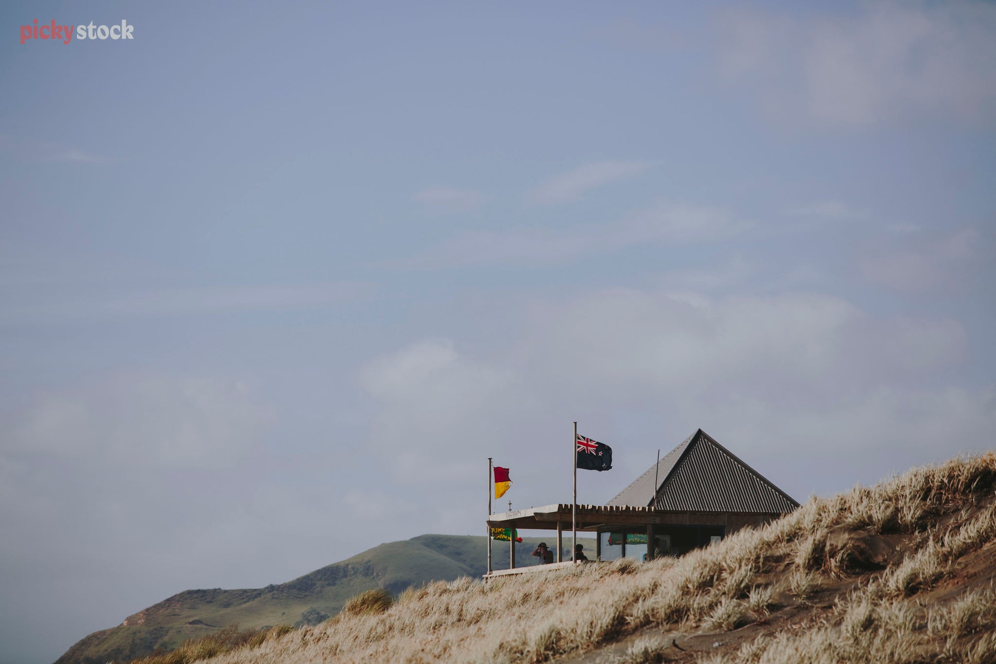 Surf club building sits nestled amongst black sand dunes on West Coast beach. The surf flags are out, seen from a side angle along the sand dunes. 
