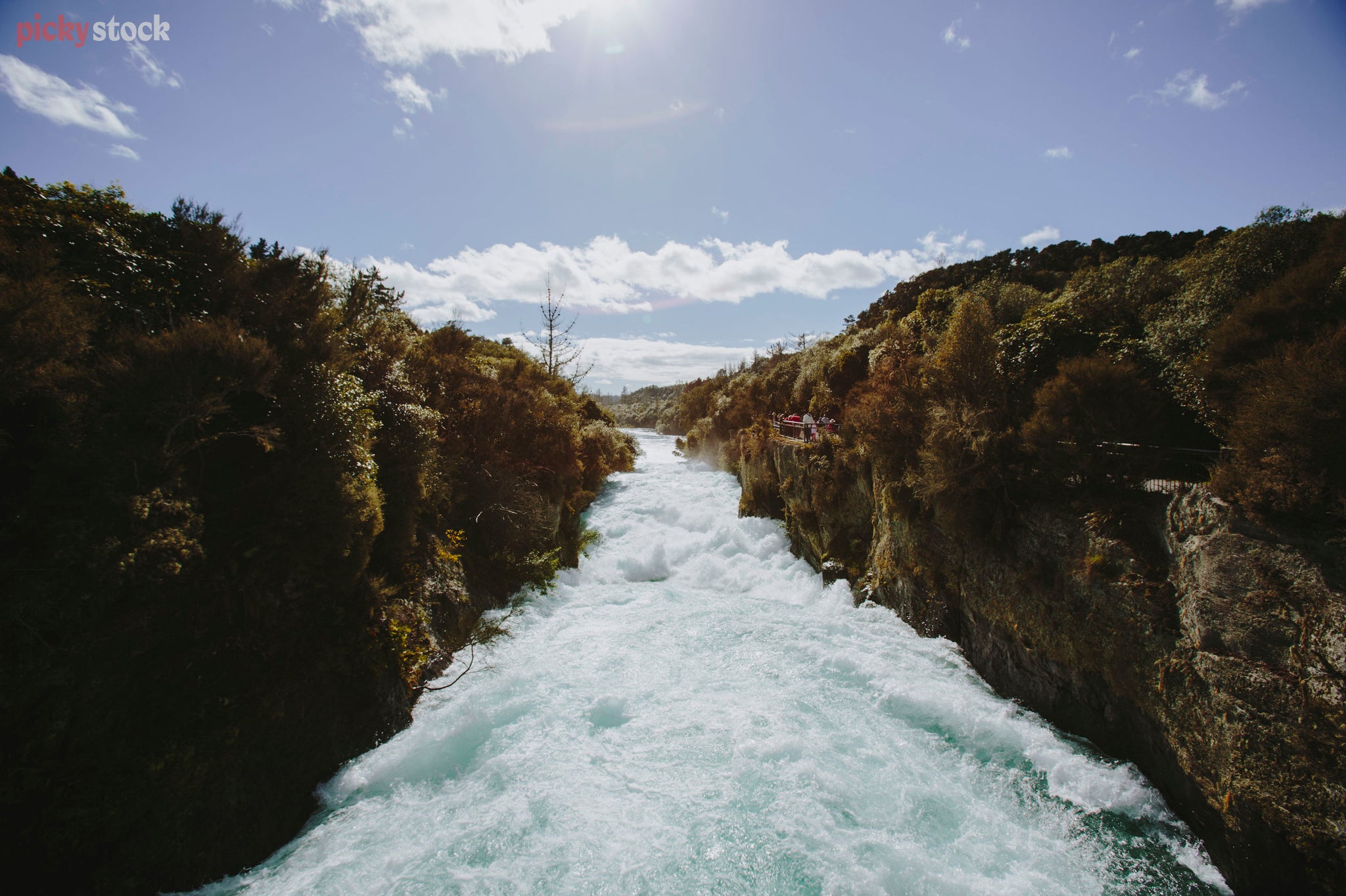 Symmetrical viewpoint from the middle of Huka Falls Bridge, the ice cold falls gushing through the middle of the image, bright blue sky above. 