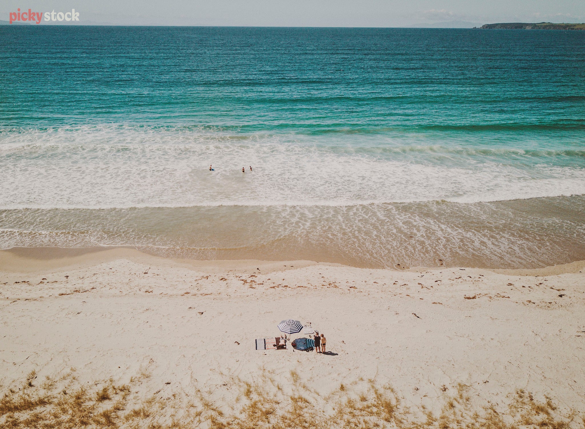 High angle aerial image of young group of people sitting on towels at the beach. The shot is very graphic, the people are small and speckled, with white sand and blue sea creating stark contrast.