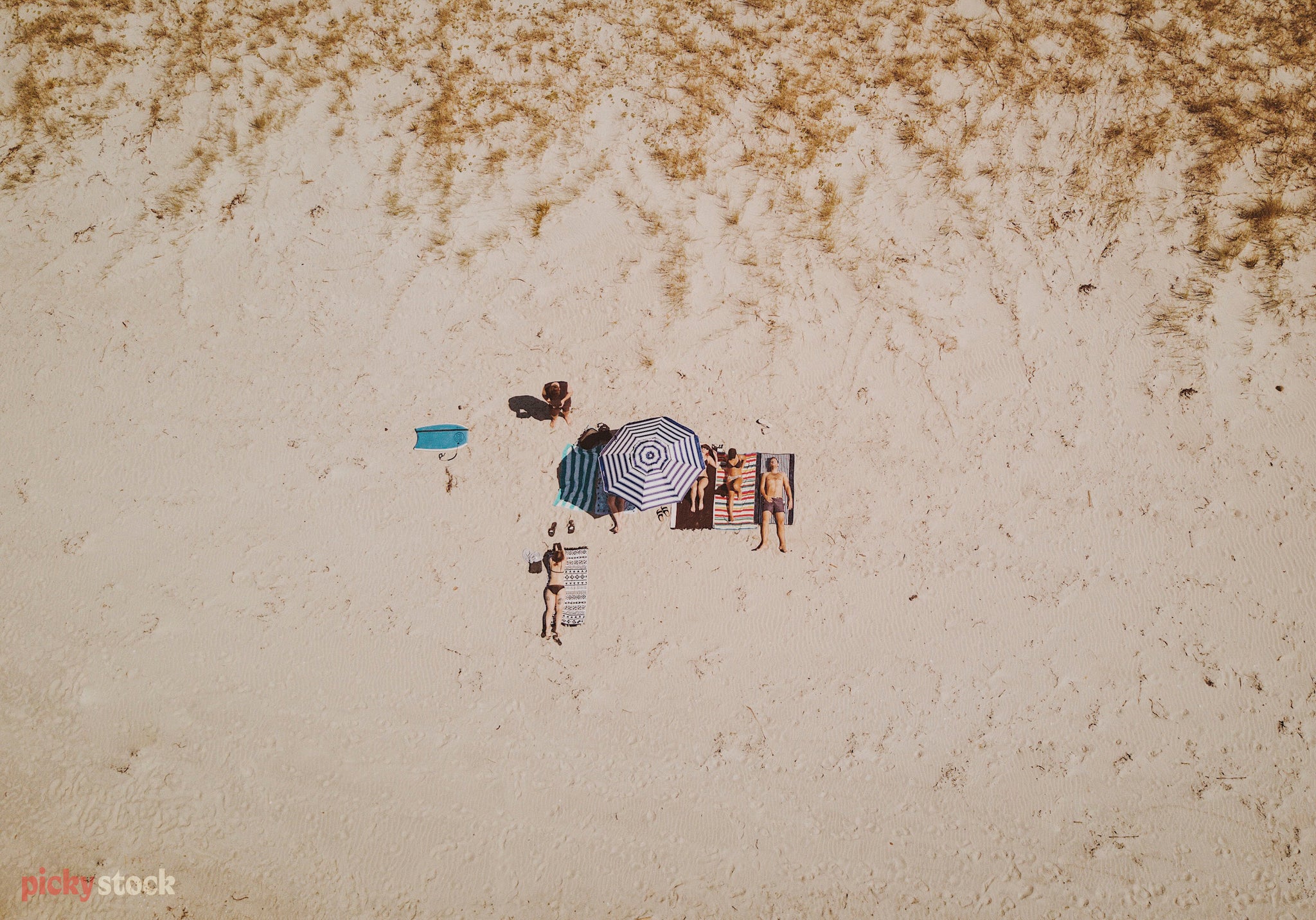 High angle aerial image of young group of people sitting on towels at the beach. The people look like colourful dots as seen from above against the white sand. 