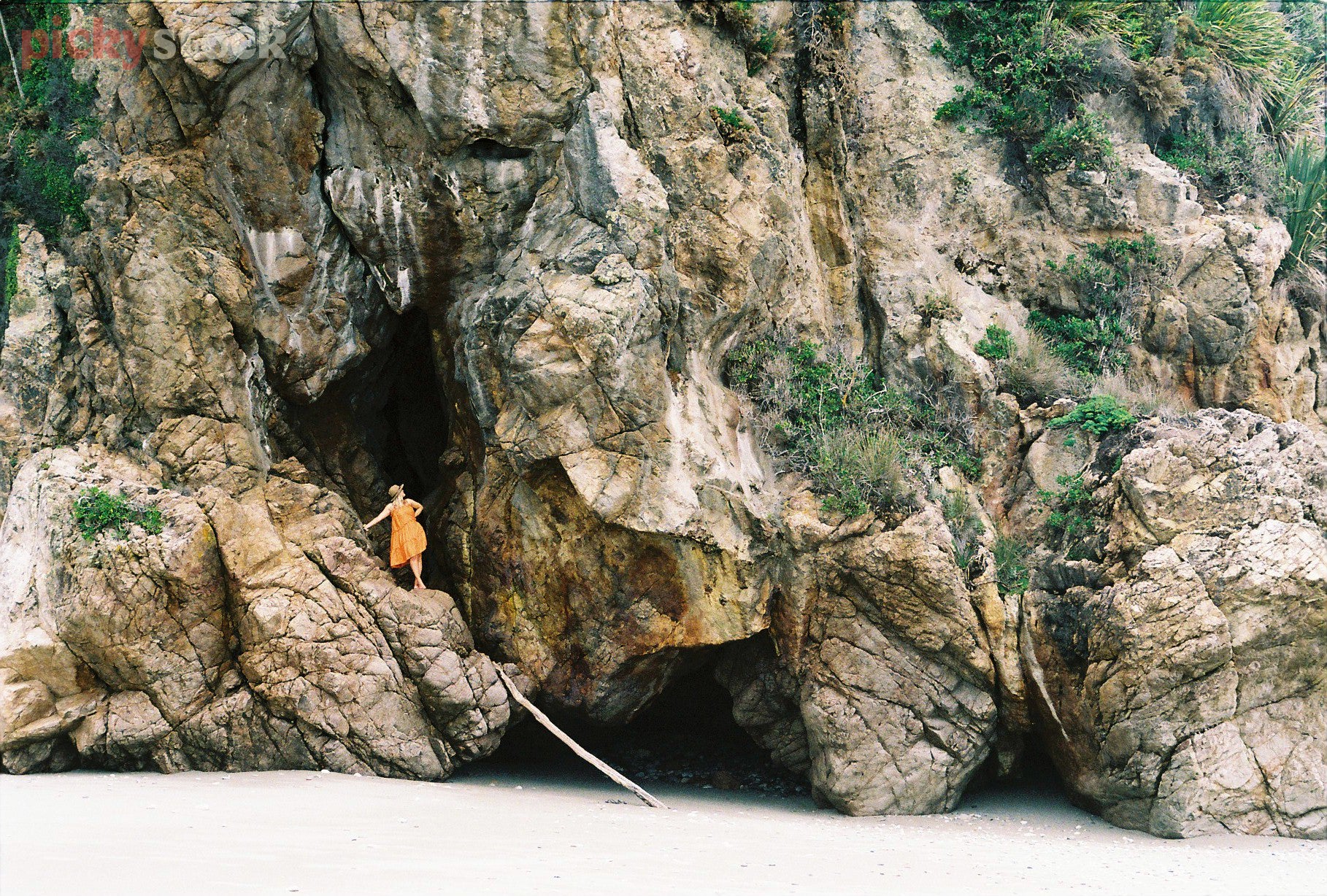 Lady with orange long dress and straw hat stands in amongst a large rock cave. She's unrecognisable, dwarfed by the rocky cliff face. 