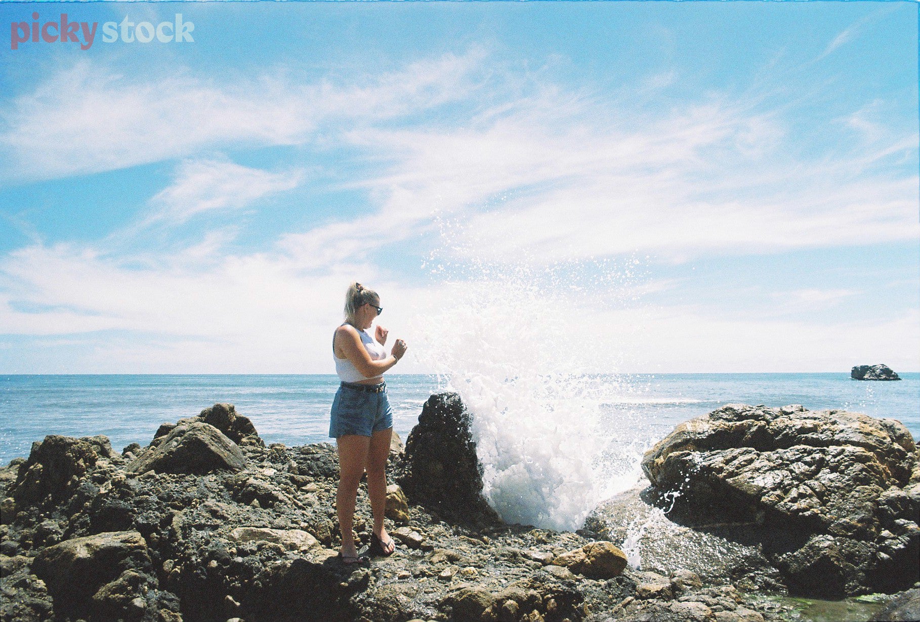 Lady stands on rocks near water, looking back over her shoulder out to sea as the salty sea spray hits her. 