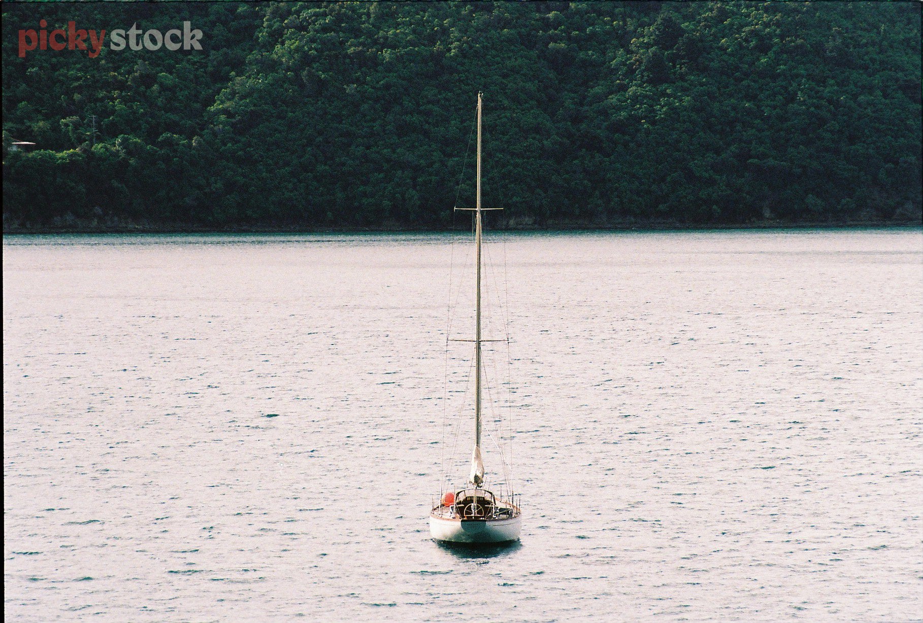 One single sailboat sits anchored in the calm bay. The boat is directed straight towards the camera, making for an angular & graphic layout. The sailboat's sail is down. 