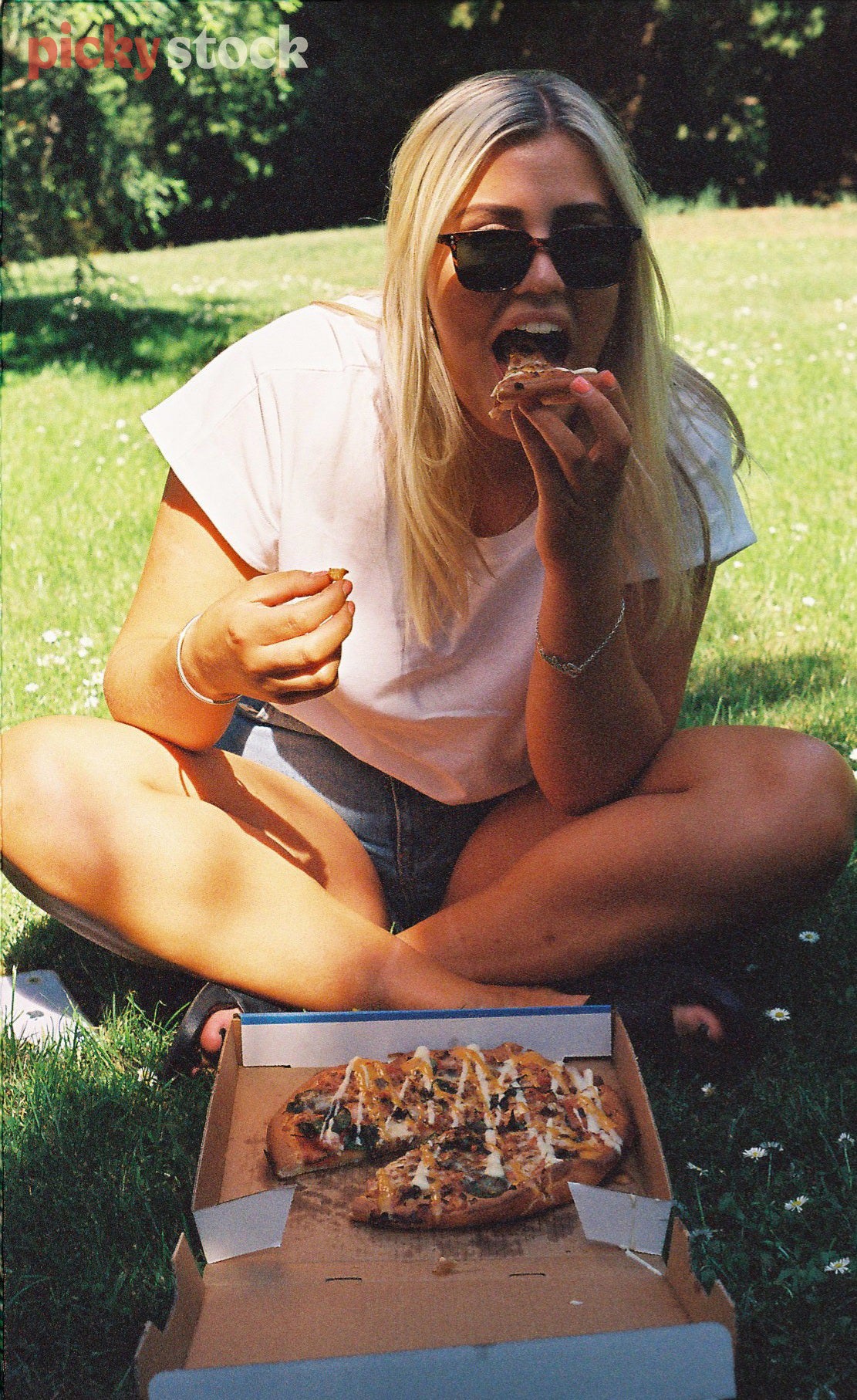 Blonde lady in her twenties bites into a small takeaway pizza while sitting on the grass in a park on a summery day. She wears a white tshirt and has denim jeans on. 