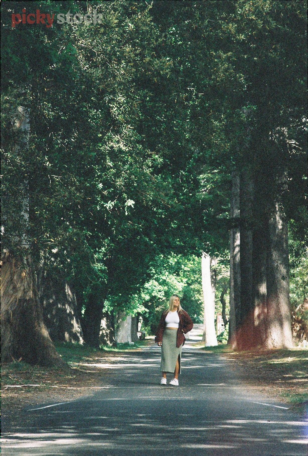Blonde lady in her twenties stands under a tree-lined path in a park. She wears a tigh white top, a longer green skirt and a brown fur jacket. 