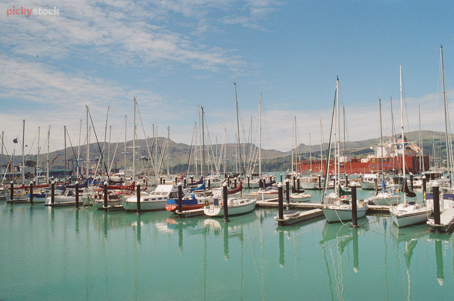 A line up of sail boats sit calmly off the docks of a marina jetty. It's a calm, blue day. 