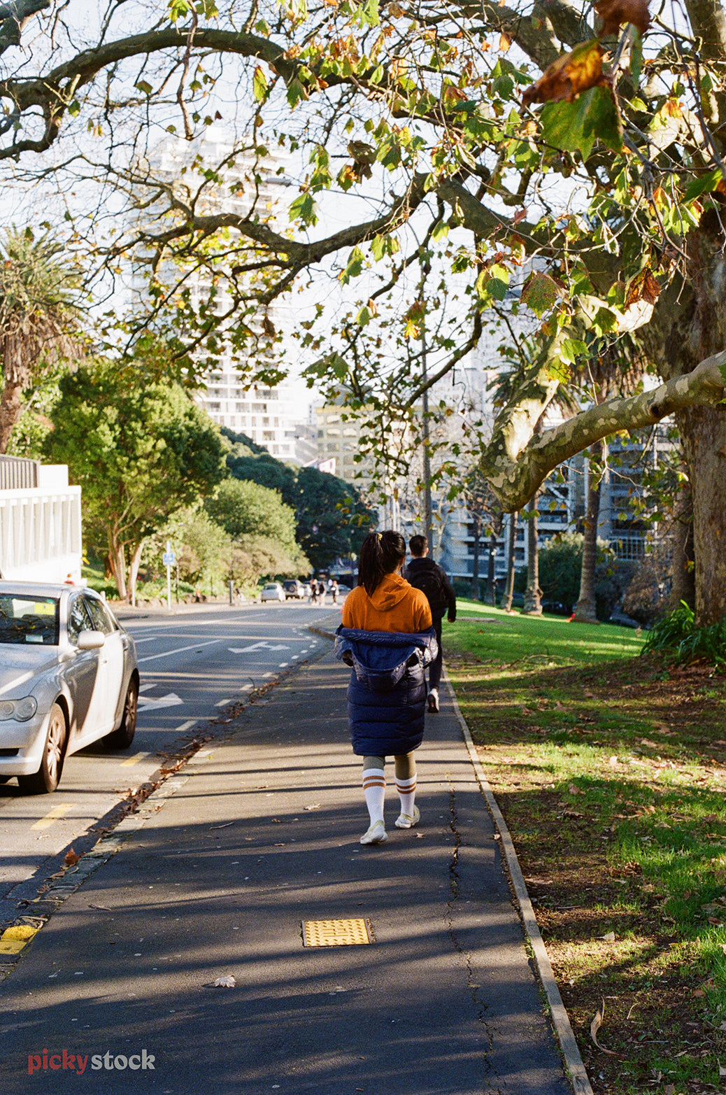 Two Asian-New Zealanders walk down the hill from Albert Park into Victoria Street in Auckland city. There's a road to the left and a park to the right. The girl has a ponytail and her windbreaker is half lowered off her shoulders. 