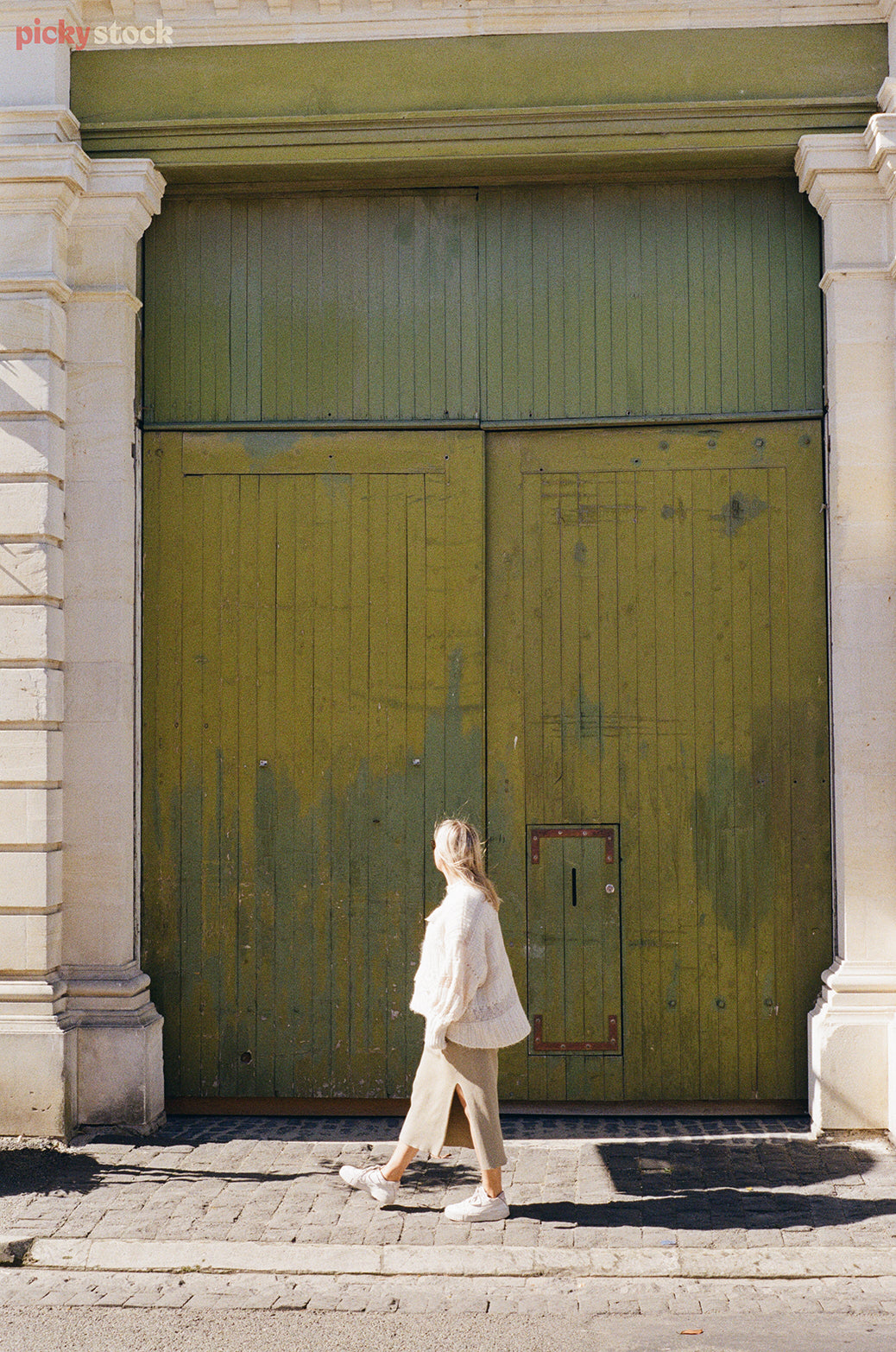 Lady with blonde hair stands in fron of large triple-head-height green washed wooden door. She's walking to the side. 
