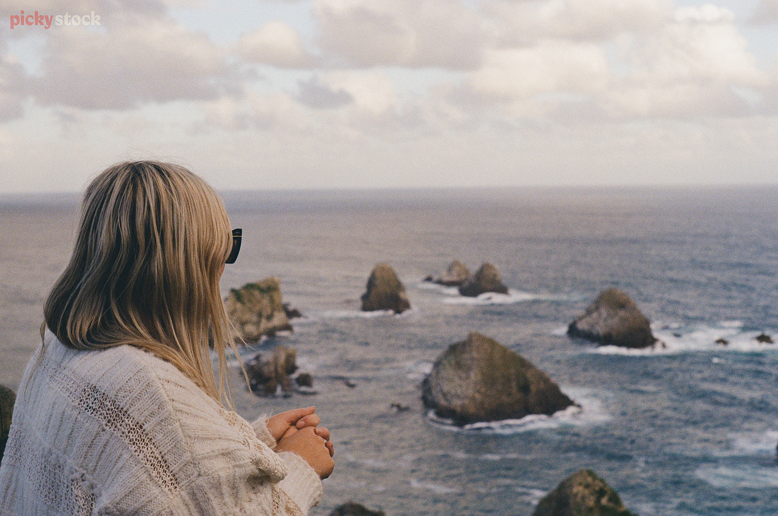 Blonde lady with sunglasses looks out over boulders in the ocean. The day is rather grey, reflected in the water. 