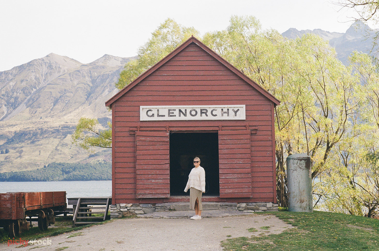 Blonde lady stands infront of the classic red weatherboard boat shed next to lake Glenorchy. 