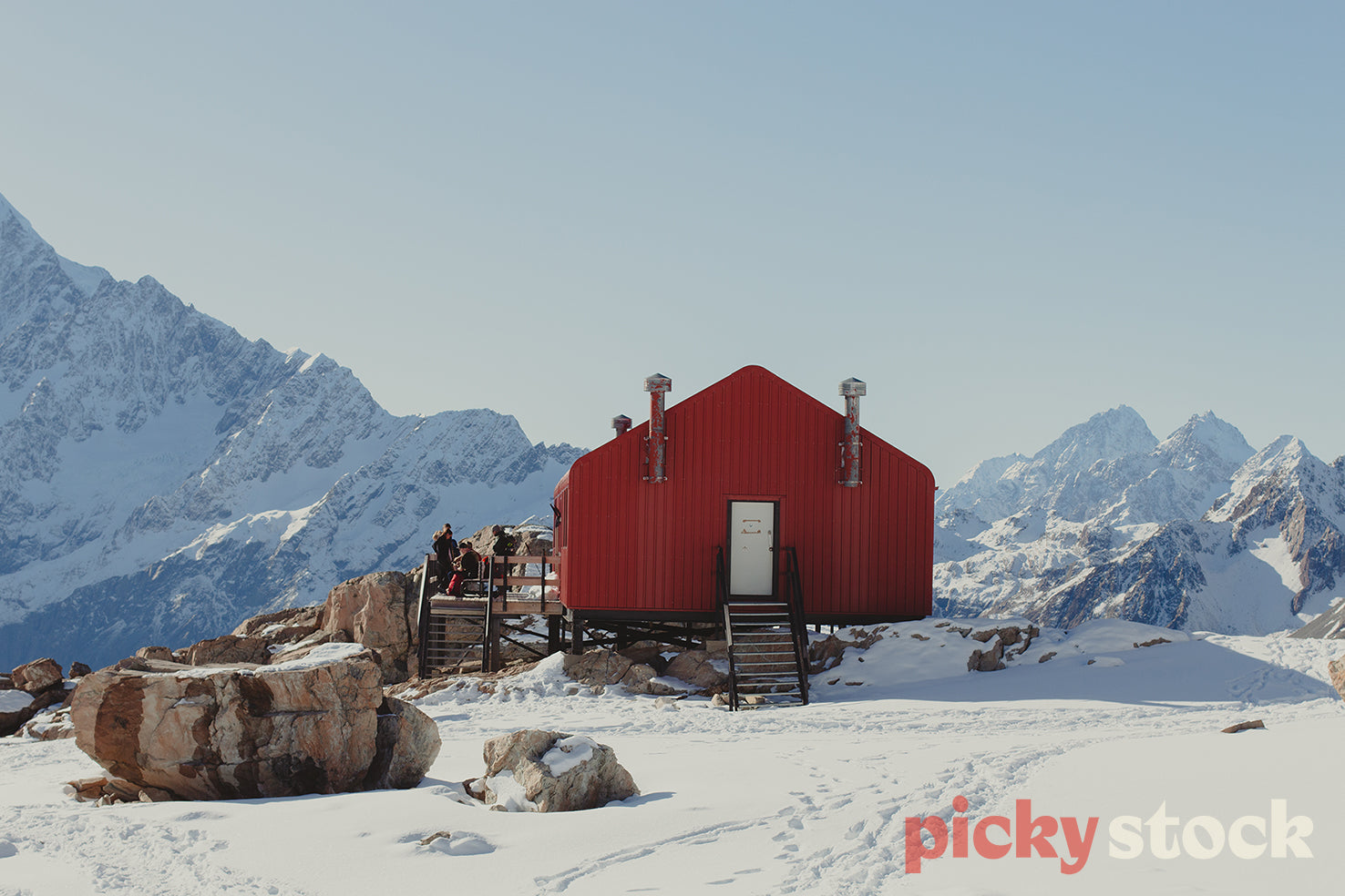 Landscape view of Muller Hut in Mount Cook during the day. Hut is red with a white door. Three people are standing outside on the deck with their tramping hiking equipment and gear. Mountains in the background covered in snow. Some rocks are exposed.