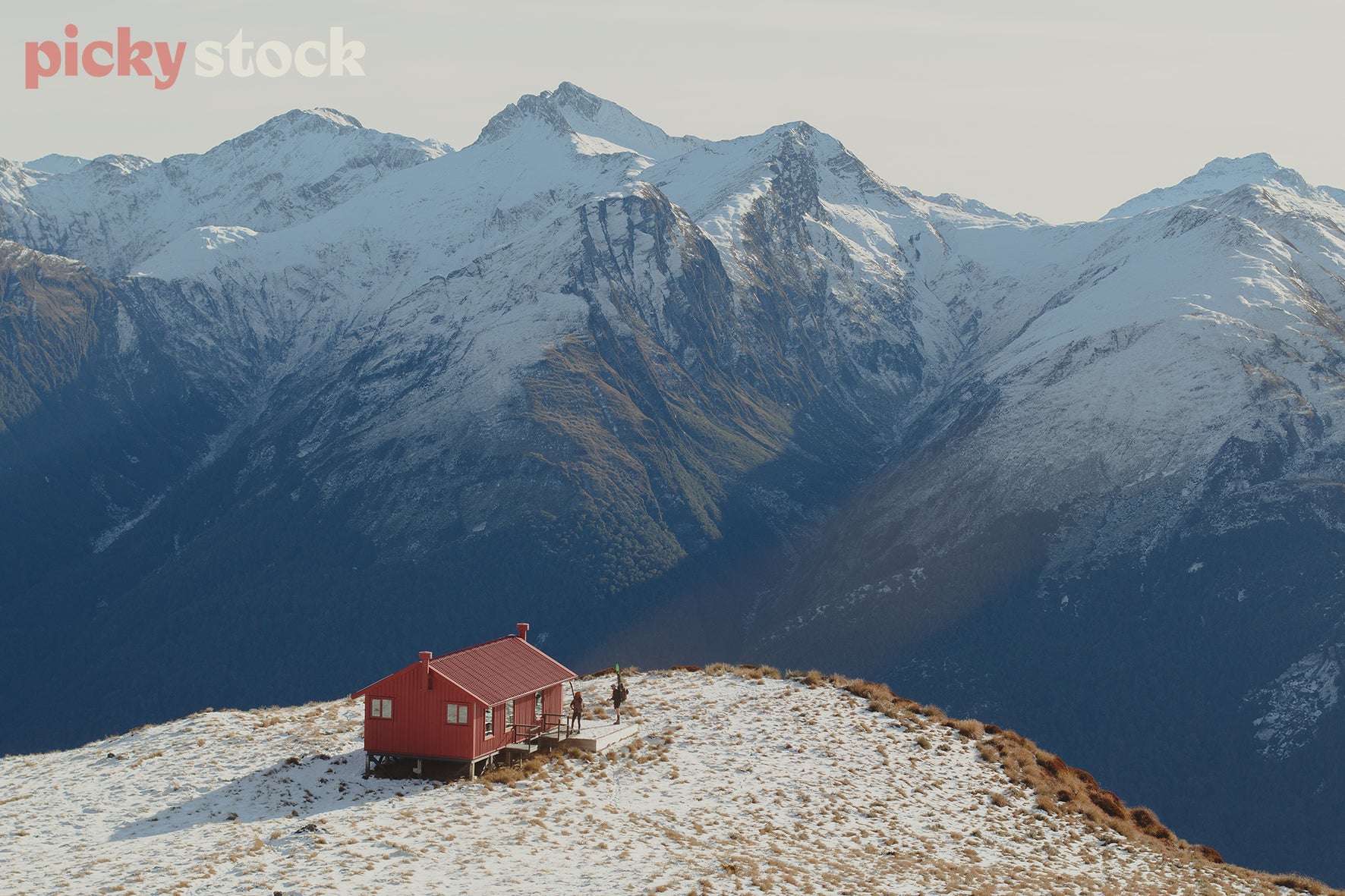 Aerial view of Brewster Hut in Mount Aspiring National Park. Snow covering the tops of the dramatic mountain range. Image captured in the middle of the day. Red hut, with a deck to the right hand side. 