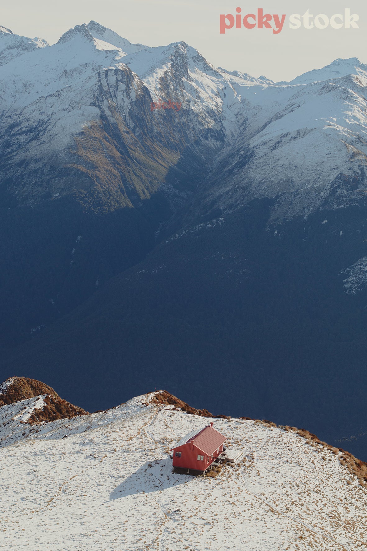 Portrait aerial image of Brewster Hut,  Mount Aspiring National Park. Hut is red, with a shadow to the left side. Low light, with dramatic snow capped mountains in the background. Hut on edge of a ridge or cliff