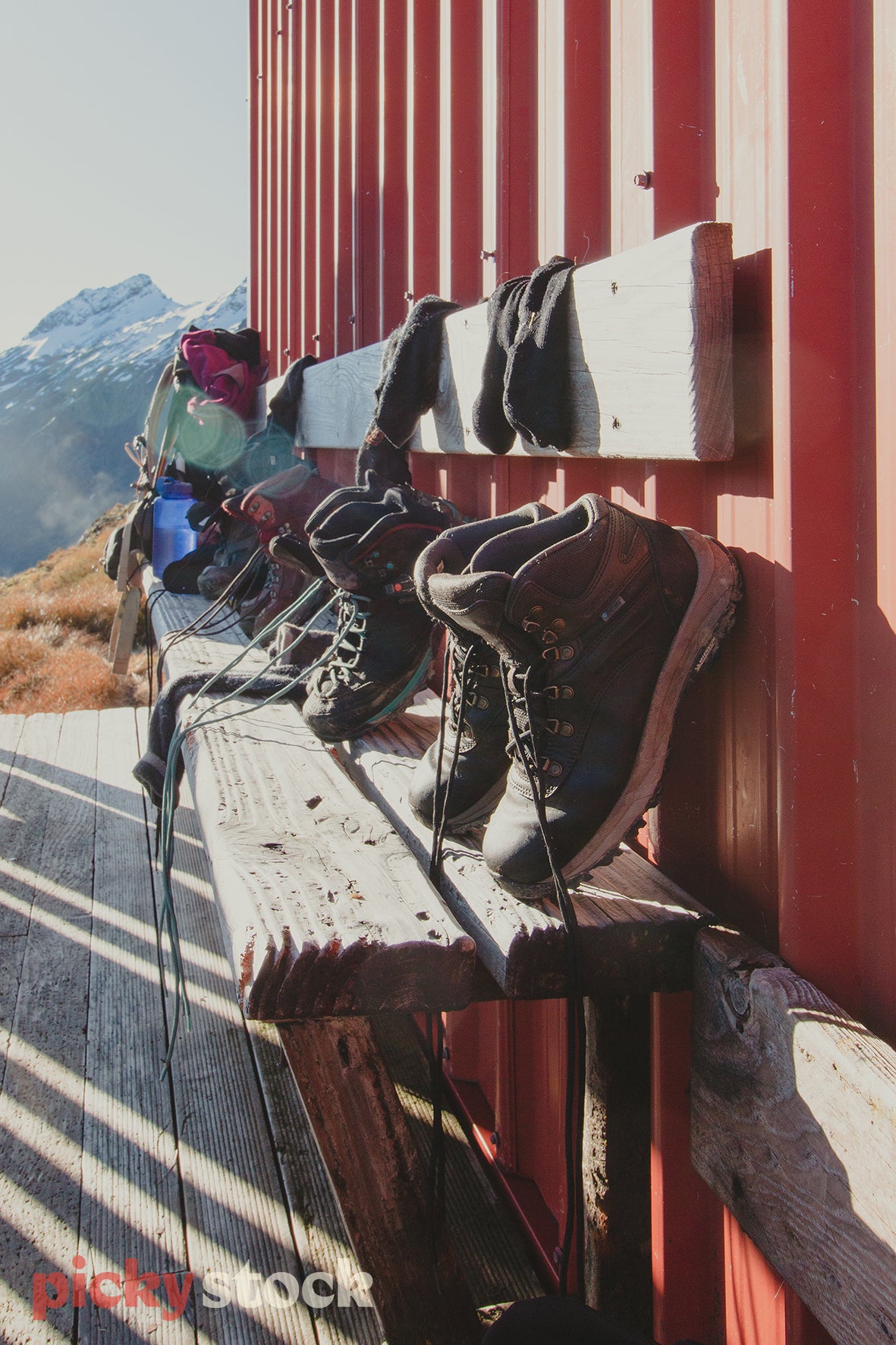 Row of tramping hiking boots outside French Ridge Hut,  Mount Aspiring National Park. Sitting on a outdoor seat, with other tramping gear at the back. Socks also drying out in the sun. Beautiful light hitting the deck creating a shadow across the image. Hut is red.