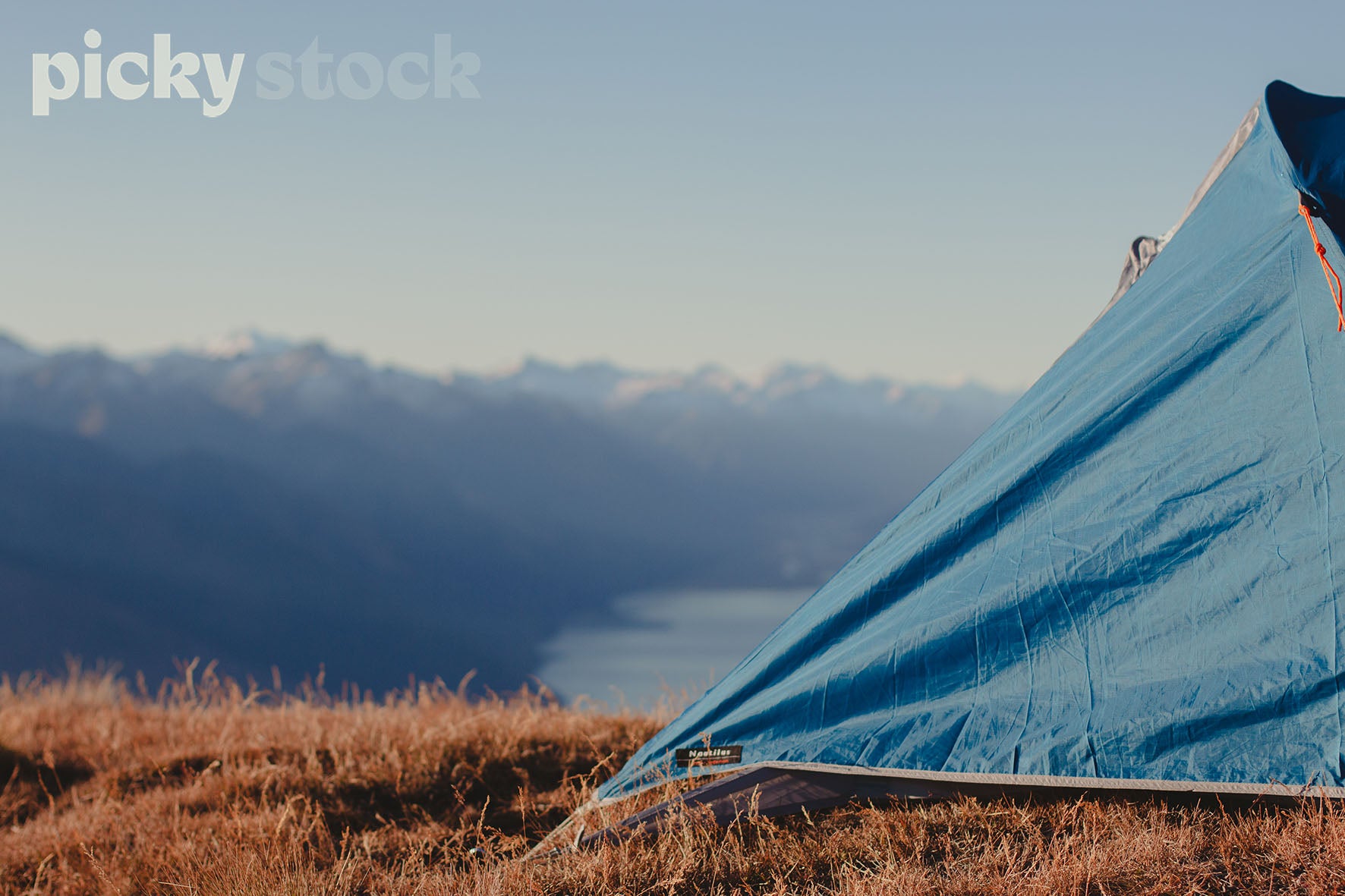 Landscape shot of part of a tent set up on dry mountain grass. View of beautiful valley, blue water and snow capped mountain range. 