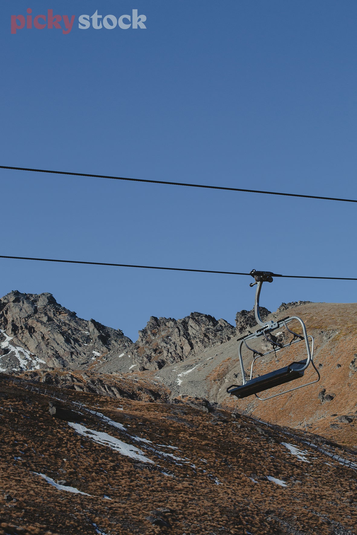 Ski field in summer. Only small patches of snow remaining. Single chair lift and cabling to the right of frame. Bright blue sky.