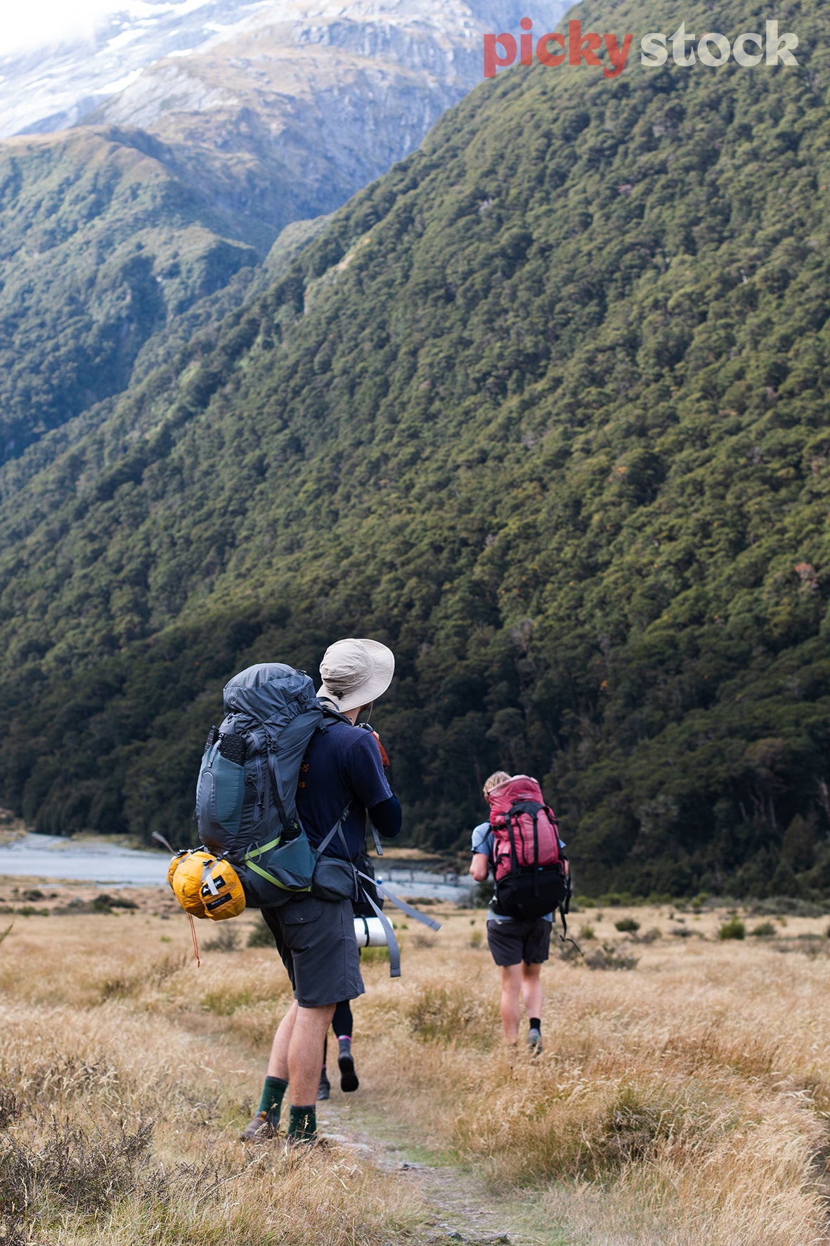 Close up of two friends walking hiking tramping through the valley of a National park in New Zealand. One Hiker closest to camera is wearing a grey bucket hat. Backpack is blue, with a yellow sleeping bag attached. They are walking through dry tussock mountain grass. You can see the river in the far background, with the large green dense mountain behind them.