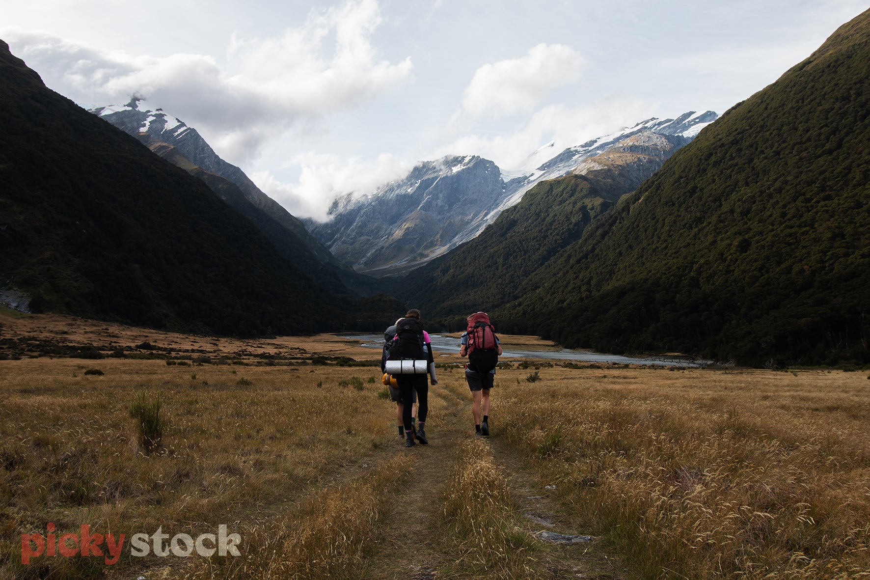 Two friends walking through the valley on a hike / tramp. Both carrying large packs, with a grey sleep mat attached. Walking through valley towards small river with mountain view, meeting in the middle of frame. Grey overcast sky.