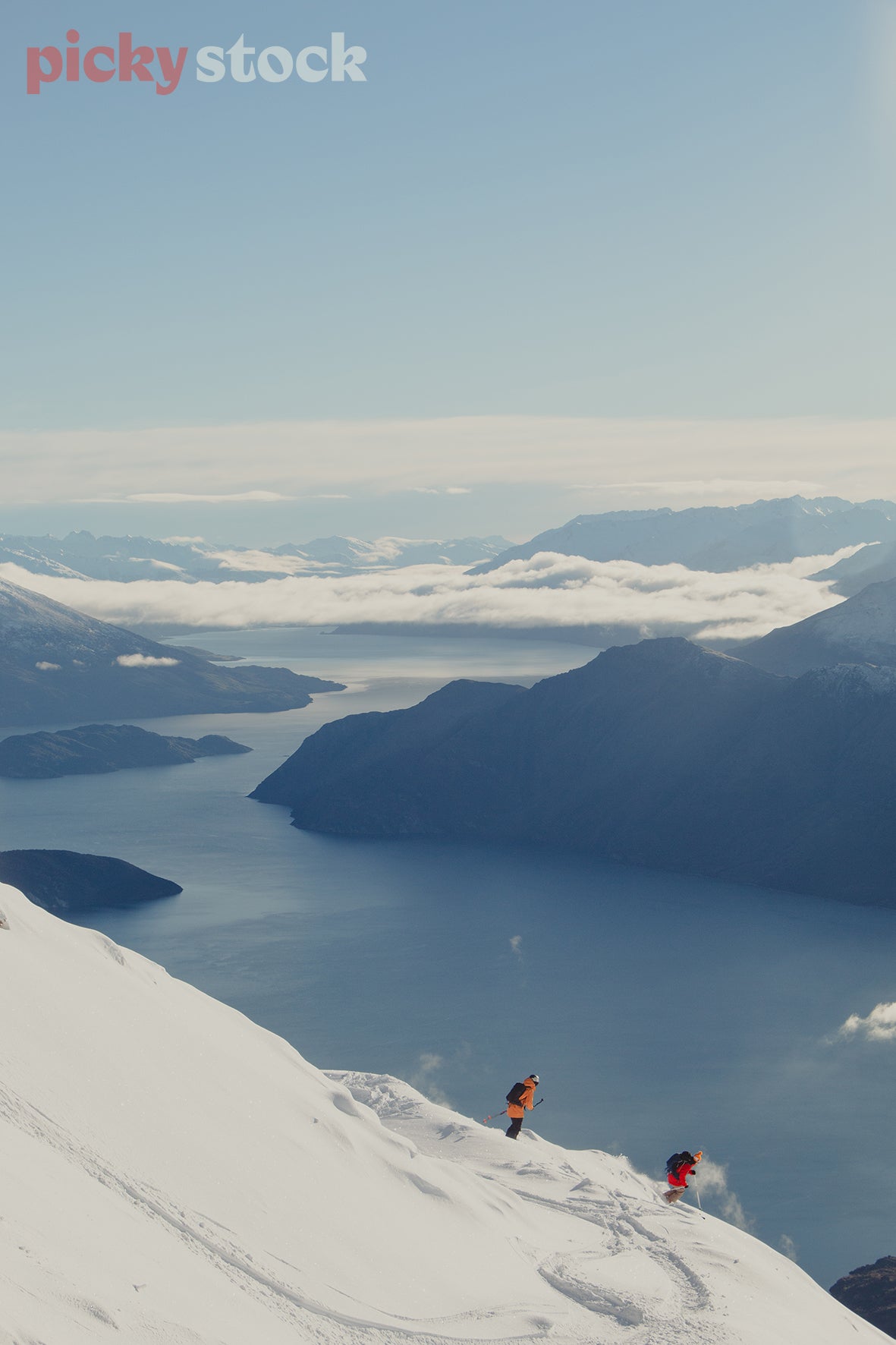 Two skiers with ski poles, skiing down the summit of Roys Peak in winter. One skier is wearing a bright orange jacket, and the other bright red. Both black backpacks, portrait view looking down at the blue body of water in Wanaka. 