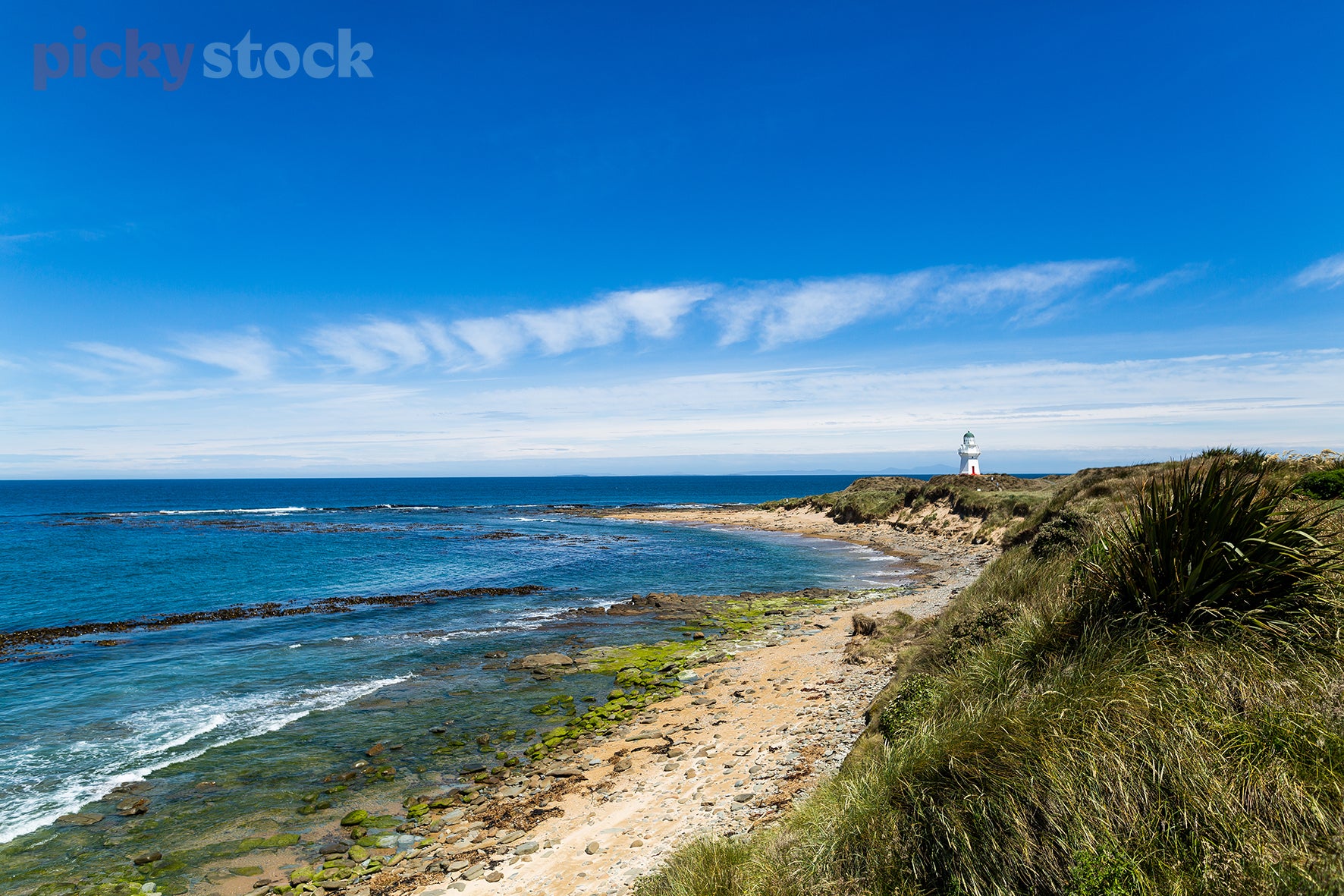 Rock beach scene with a lighthouse in the far distance. Sky is very blue with scattered cloud.  Water is blue and calm. 