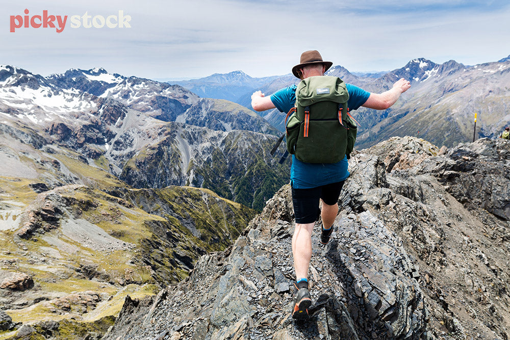 Man walking along dramatic rock formation and mountain range. Wearing large green backpack. Wearing hat. 