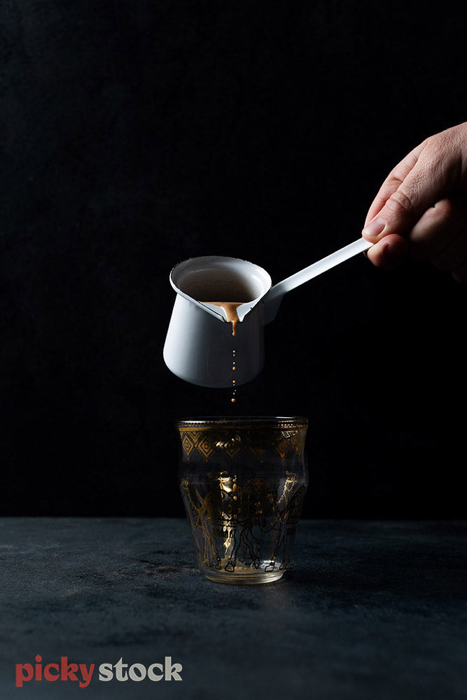 Dark moody shot of hand holding small jug pouring a hot dark drink. Pouring into Moroccan style glass. Shot is capturing the image mid pour