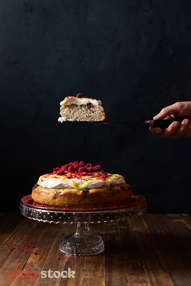 Hand in frame picking up slice of cake from cake stand. Background is dark. Cake on wooden surface. Cake has a pile of fresh raspberries and a thick white icing with bits of lemon rind.  
