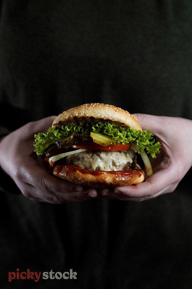 Hands in black t-shirt and dark background holding large burger. Sesame bun with fancy lettuc, tomato, pickle, melted cheese on thick beef pattie