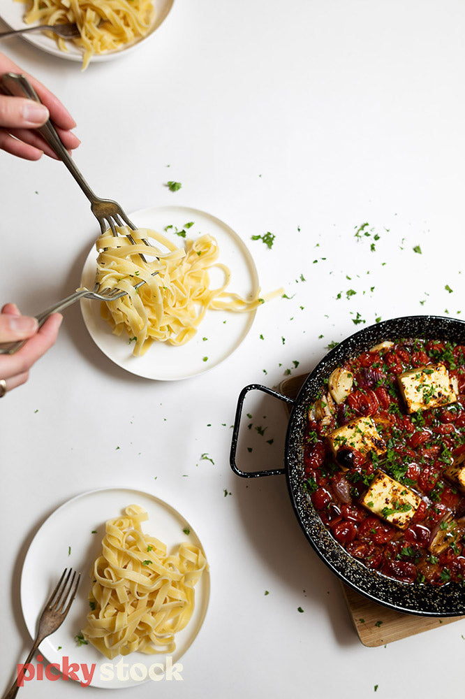 Italian pasta dish being served on white table setting. Herbs scattered off the plated areas. Forks in frame picking up fresh pasta. Large cast iron pan filled with thick tomato and halloumi sauce. 