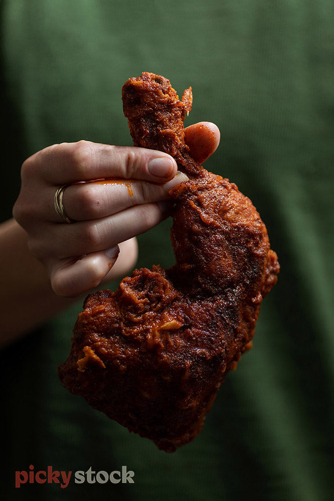 Female hand wearing ring, wearing green top. Holding cooked coated chicken wing. 