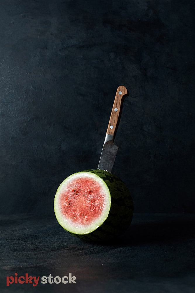 Portrait image of cut watermelon with large knife with wooden handle inserted in back of melon. Background is a dark black texture. Mood lighting