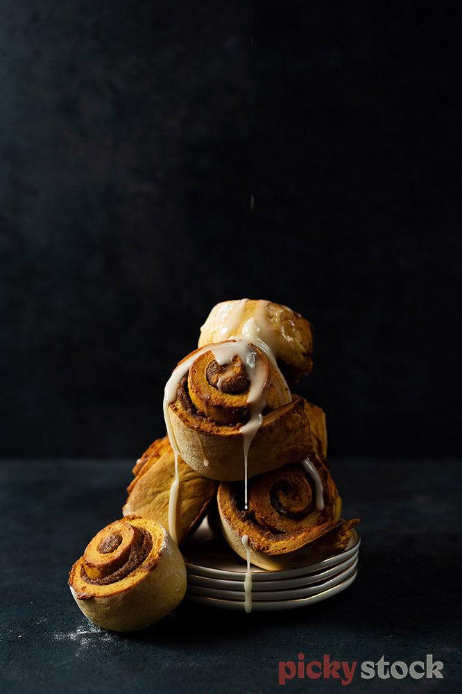 Freshly cooked cinnamon buns stacked up on a bunch of plates. Icing dripping down onto surface. Dark black background.