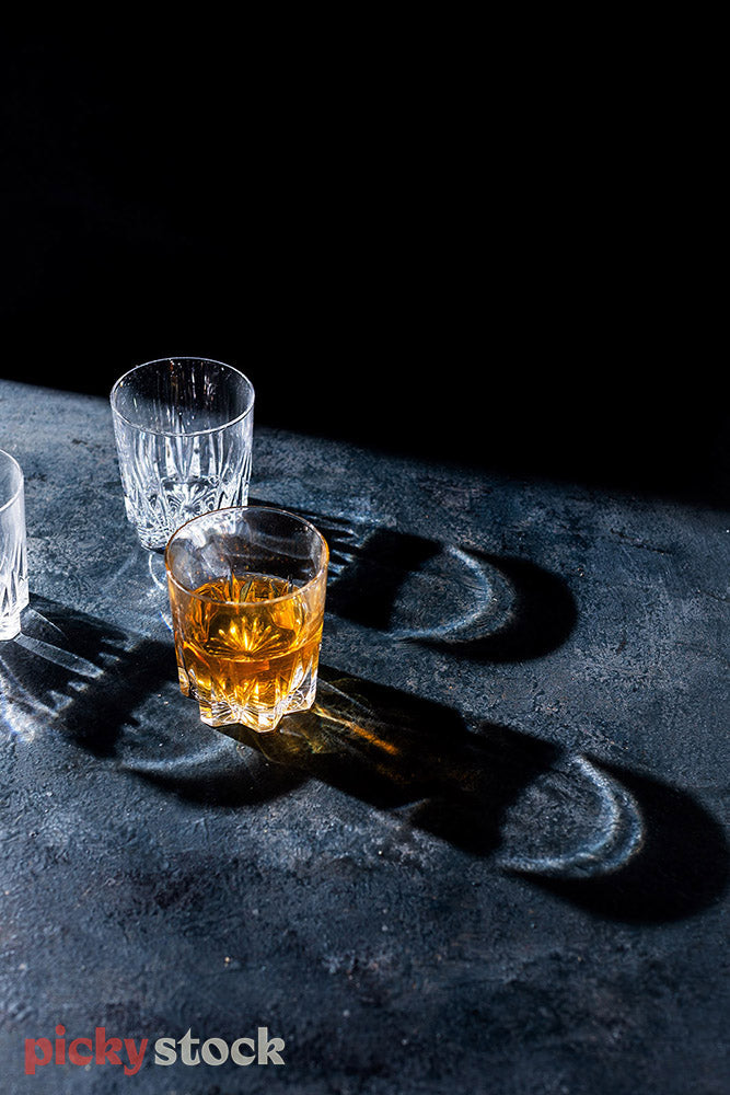 Three glasses on marble surface. One drink filled with brown liquor. Marble surface with dark black background.