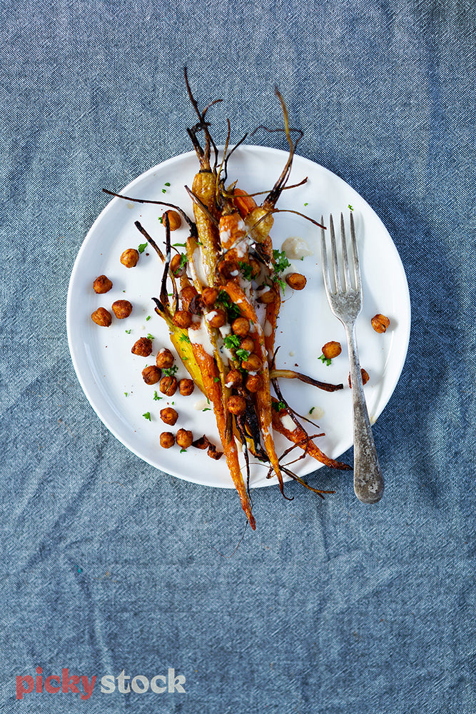 Delicious roast carrot and chickpea dish with yoghurt dressing sitting on white plate and denim blue tablecloth