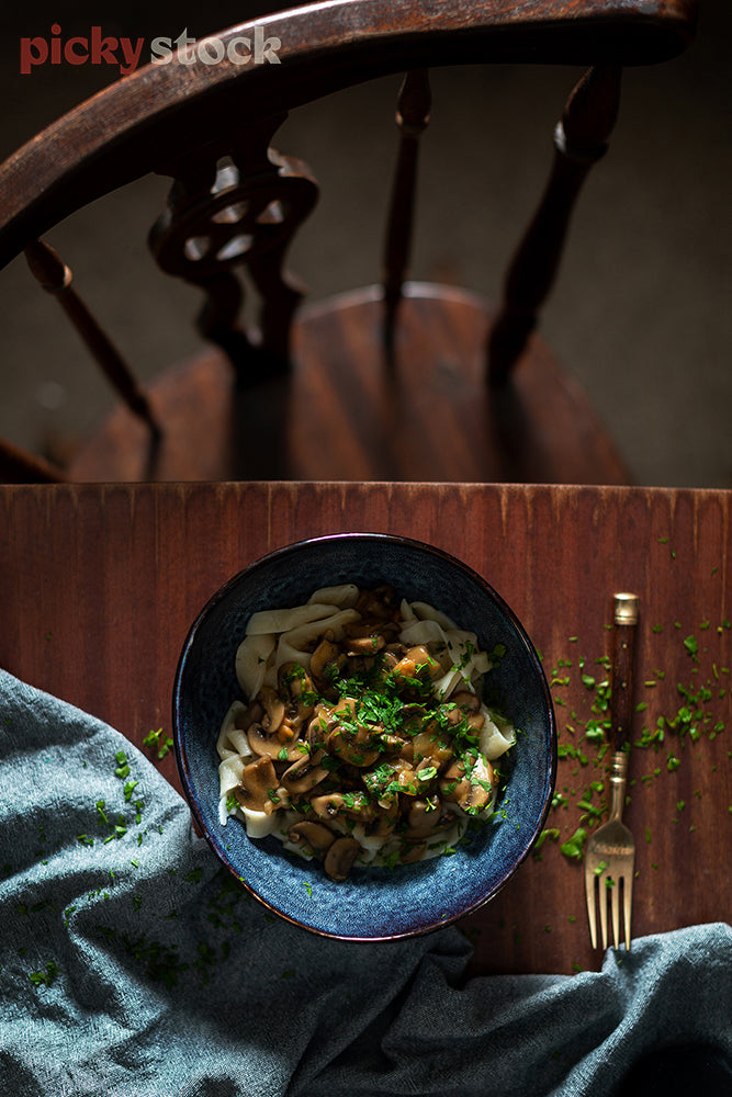 Mushroom pasta dish in dark bowl sitting on dining table with empty chair. 