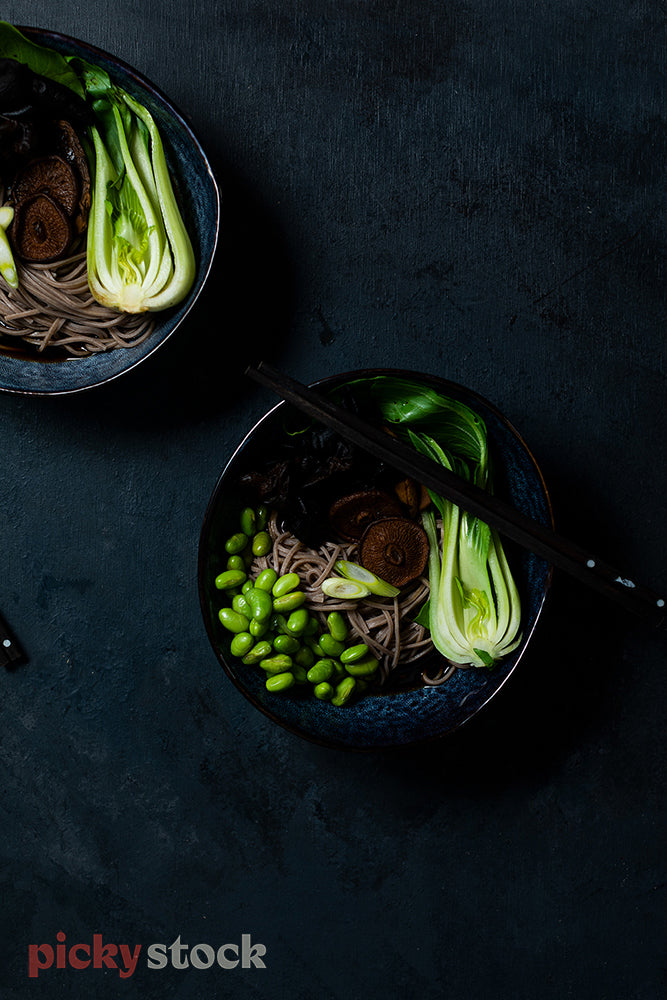 Two fresh bowls of food against dark background with chop sticks. Edamame, bok choy and noodles. 